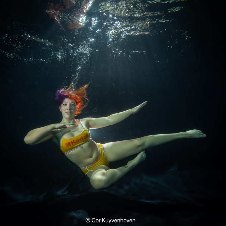Duikfoto van een vrouwelijke sporter in een donkere wateromgeving, gekleed in een felgeel badpak met een opvallende "Kodak" logo, met kleurrijk haar en een krachtige pose.