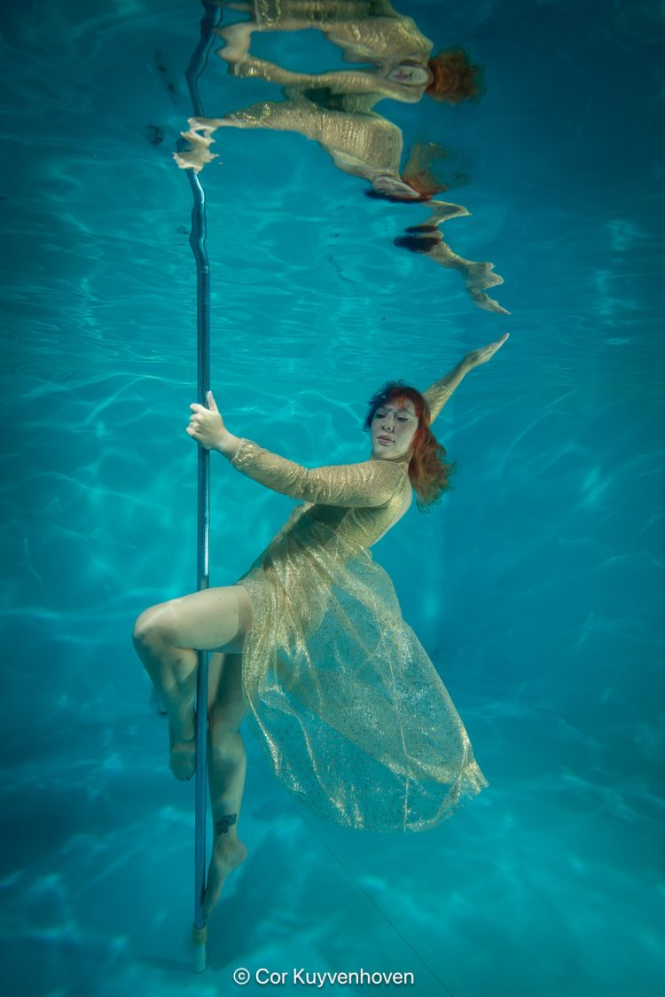 Alternatieve onderwater foto van een vrouw in een gouden jurk die balanceert met een paaldans paal in een zwembad.