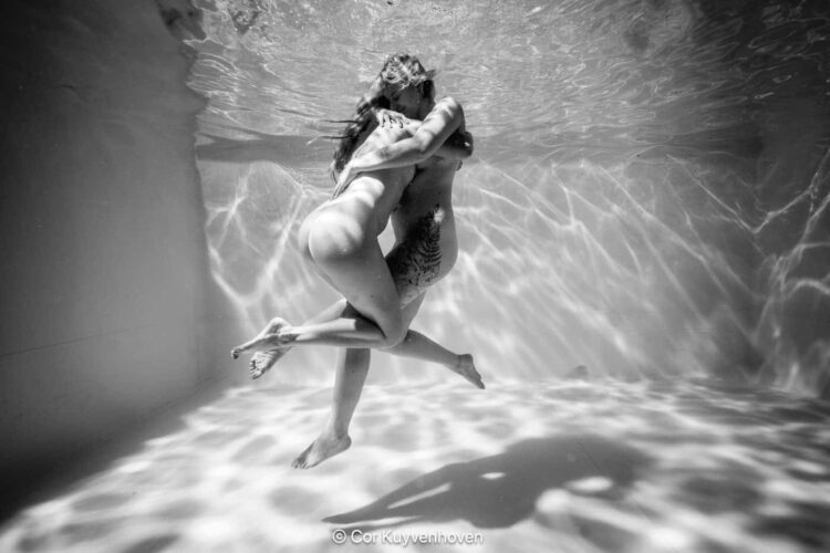 Onderwater kus tussen twee vrouwen in helder zwembad, zwart-wit sfeer, gevoel van verbondenheid en liefde, artistieke fotografiestijl.